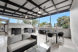 A modern outdoor patio with a sofa, dining table, chairs, ceiling fans, and large windows offering a view of trees and buildings.