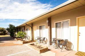 Um motel com área de estar coberta ao ar livre, estacionamento e um vaso de tijolos com uma pequena árvore.