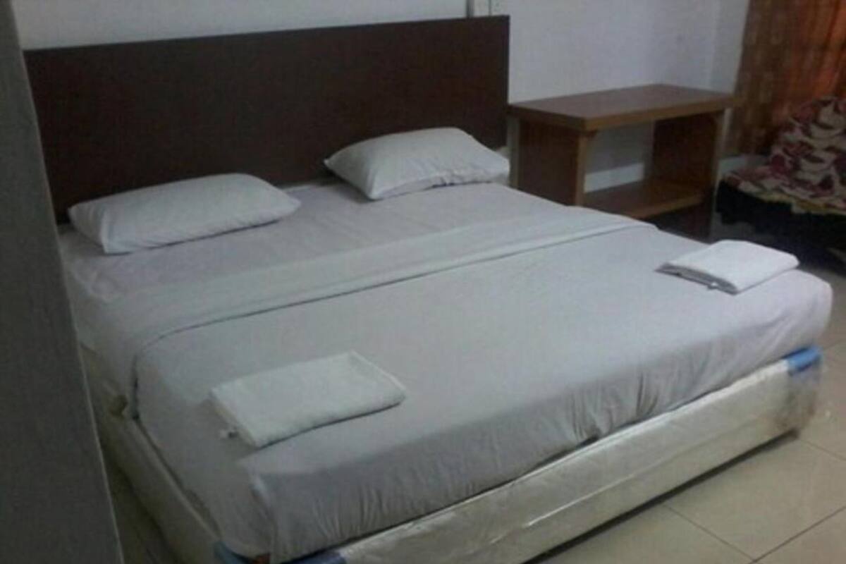 Una cama individual con ropa blanca, cabecera de madera, mesita de noche de madera pequeña y suelo de baldosas.
