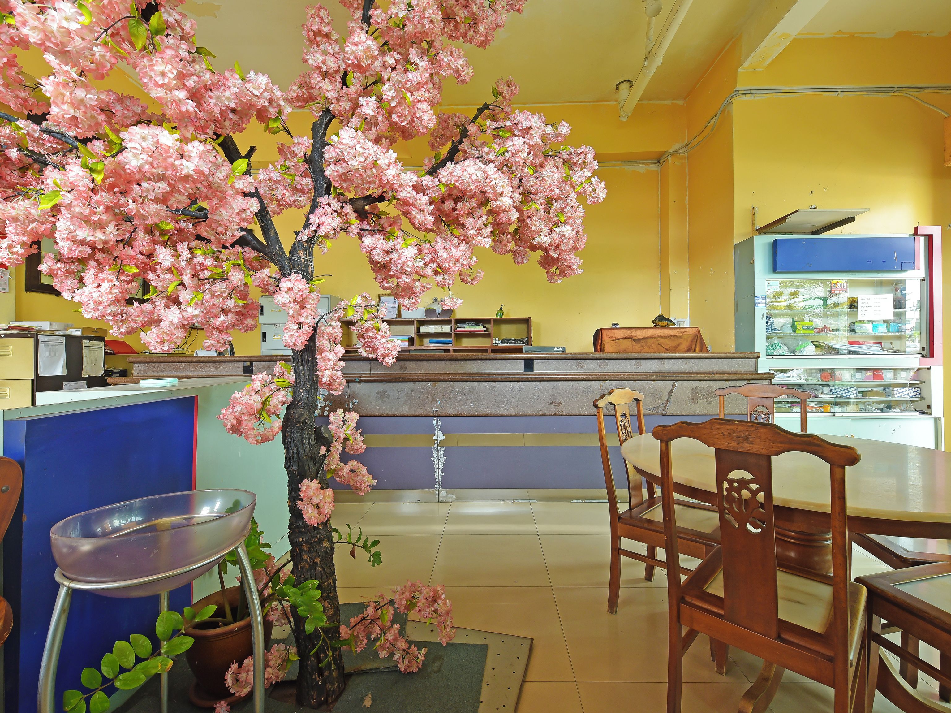 A restaurant interior with a central counter, wooden chairs, a potted tree with pink blossoms, and a tiled floor.