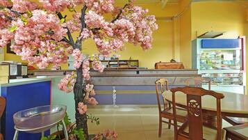 A restaurant interior with a central counter, wooden chairs, a potted tree with pink blossoms, and a tiled floor.