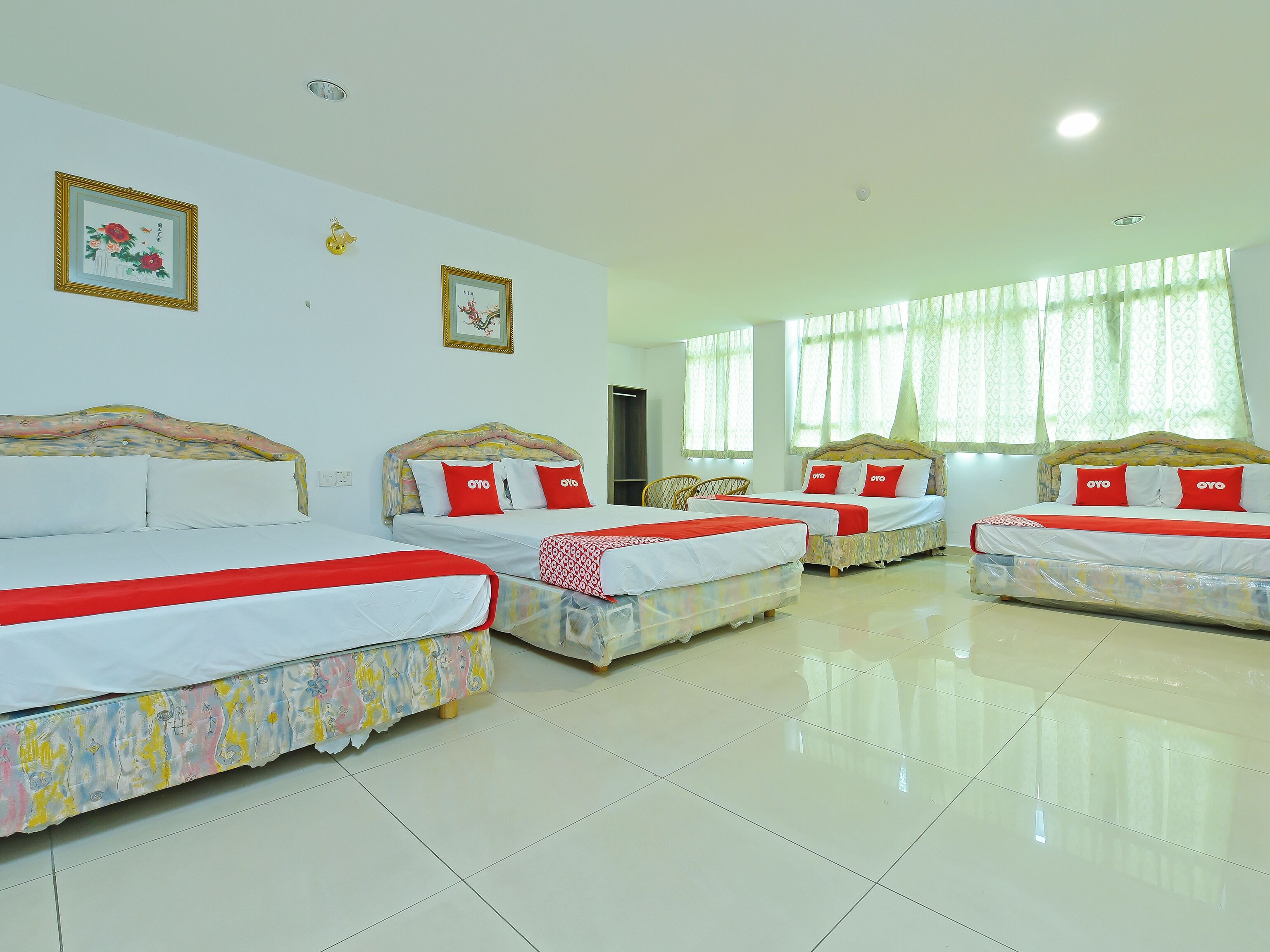 A hotel room with four beds, each with a red and white bedspread, and a large window with sheer curtains.