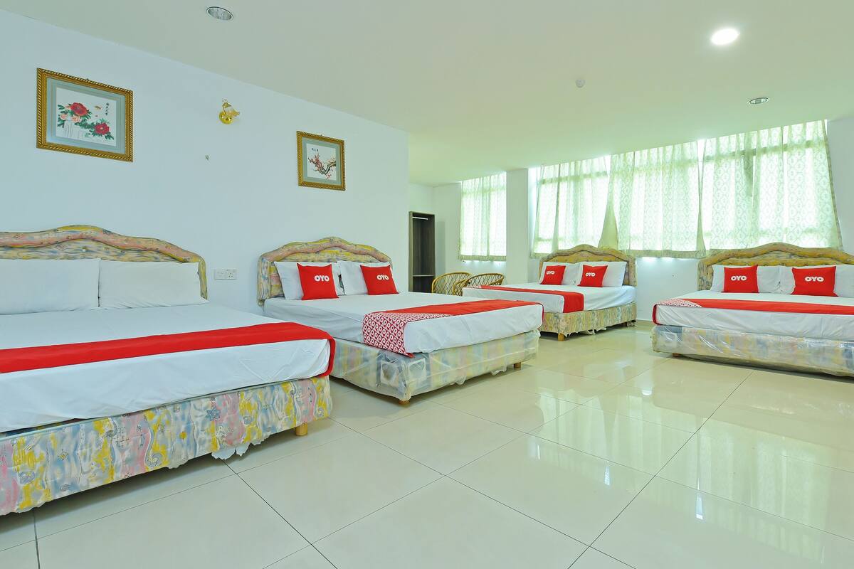 Habitación de hotel con cuatro camas, cada una con cubrecama rojo y blanco, y un ventanal con cortinas translúcidas.