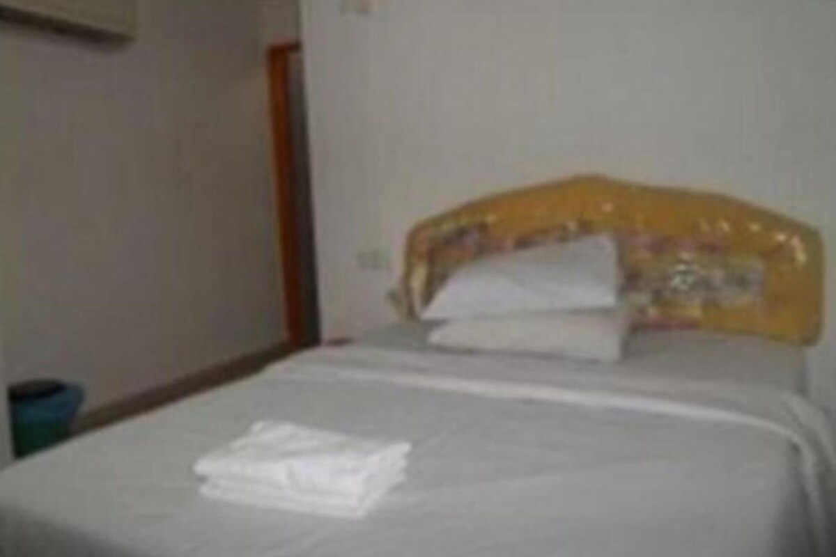 A bed with a white mattress and a yellow headboard, a folded white towel on the bed, and a blue trash bin to the left.