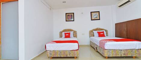 A hotel room with two beds, each with a patterned bedspread and a red pillow. There is a ceiling fan, an air conditioning unit, and two framed pictures on the wall.