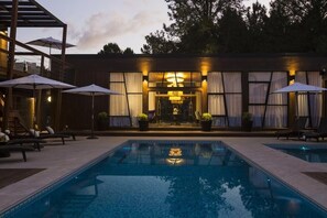 Una piscina con sillas de descanso y sombrillas, un edificio con ventanas y cortinas grandes, y una entrada bien iluminada.
