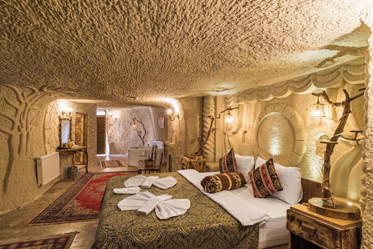 Ein Zimmer mit Steindecke, einem Bett mit weißen Leinen, einem roten Teppich mit Muster und einem Nachttisch aus Holz mit Lampe.