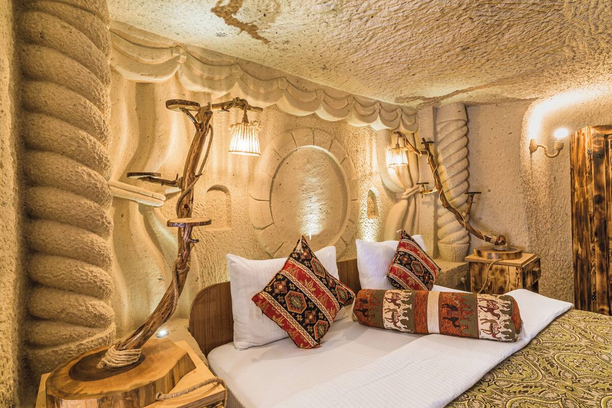 Une pièce avec un mur en pierre, un lit en bois agrémenté d’oreillers à motifs et une lampe de sol en bois au design original.