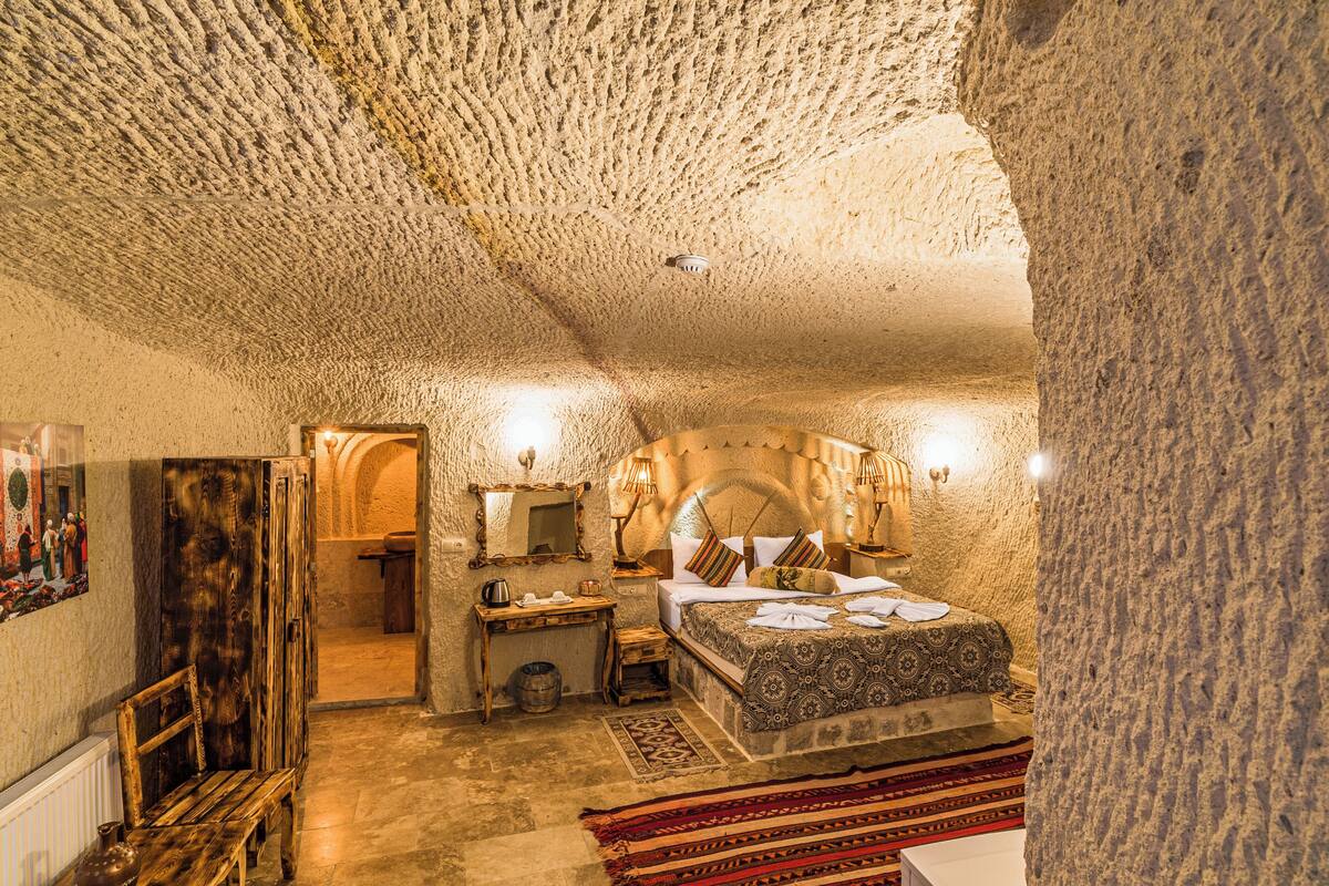 Ein Zimmer mit Steindecke, einem Bett mit gemusterten Textilien, einer hölzernen Bank und einem kleinen Tisch mit Wasserkocher.