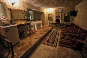 A rustic stone-walled room with a fireplace, a wooden sofa, a dining table with chairs, and a kitchen area with a refrigerator and kettle. - Cappadocia Ennar Cave and Swimming Pool (Nevşehir)