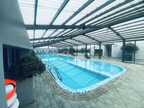An indoor swimming pool with clear blue water, surrounded by lounge chairs and a covered area with a metal roof. - Lancaster Luminaire - Hoba Housing (Hà Nội)