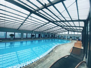 Ein Hallenbad mit Glasdecke, klarem Wasser und Liegestühlen am Rand.