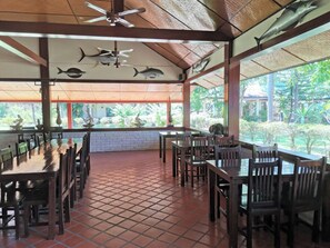 A covered dining area with tiled flooring, wooden tables and chairs, ceiling fans, and fish wall art.