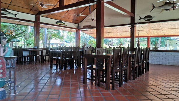 A spacious indoor dining area with high ceilings, wooden beams, and large windows overlooking greenery.