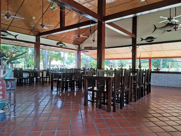 A spacious indoor dining area with high ceilings, wooden beams, and large windows overlooking greenery.