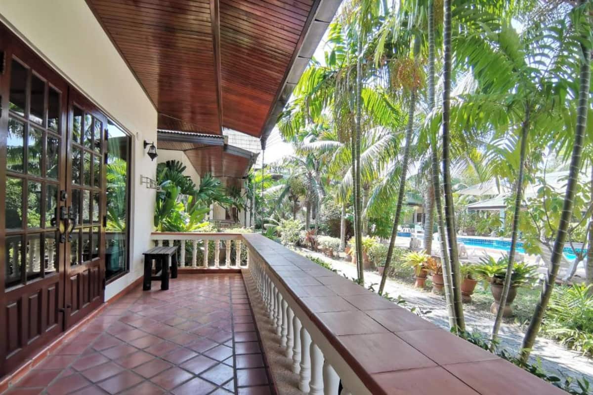 Un espace extérieur abrité, doté d’un sol carrelé, d’une rambarde en bois et offrant une vue sur une piscine et des palmiers.