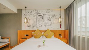 Habitación de hotel con cama grande, cabecera de madera, dos mesitas de noche, una mesita auxiliar, un escritorio pequeño, una silla, una ventana con cortinas y un mural en la pared.