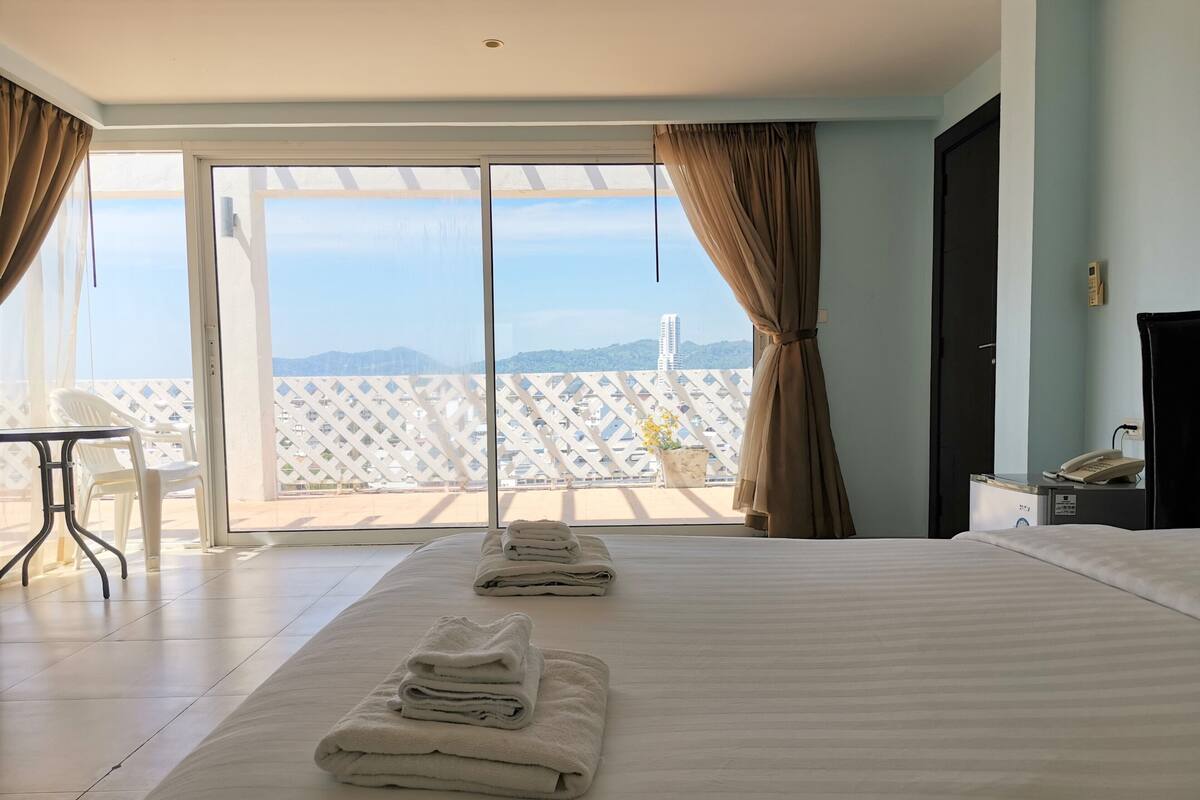 A hotel room with a large bed, a desk, and a view of the cityscape through the balcony.