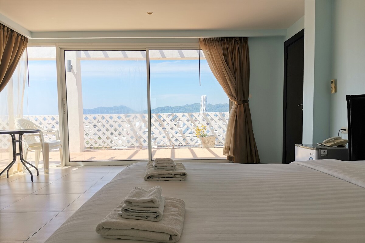 A hotel room with a large bed, a desk, and a view of the cityscape through the balcony.