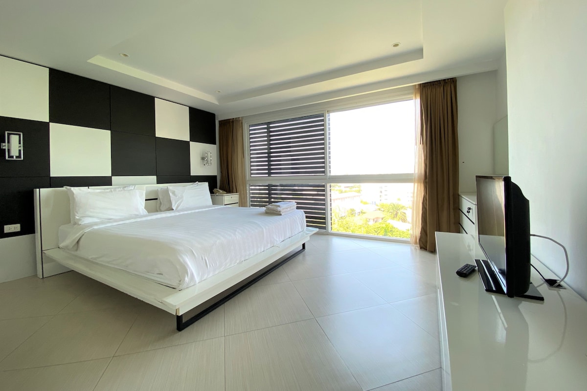 A modern hotel room with a large bed, a flat-screen TV, and a view of greenery outside.