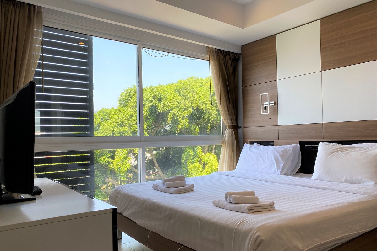 A modern hotel room with a large bed, a desk, and a view of trees through the window.