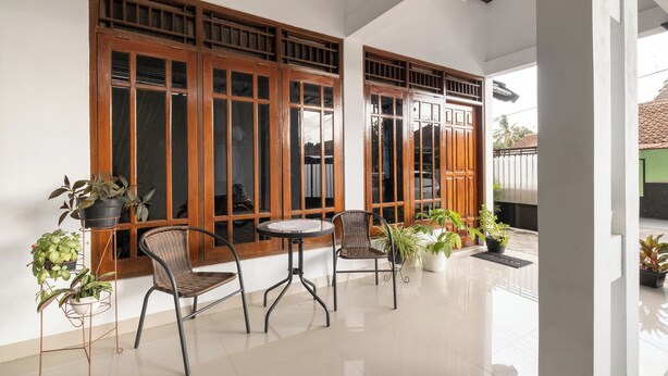 A room with a tiled floor, two wooden-framed windows, a round table, two wicker chairs, and potted plants. - Oyo 2392 Kost Safira (Ngaglik)