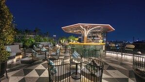 Un bar en la azotea con suelo de baldosas geométricas, asientos exteriores con barandillas de metal y una zona de bar con toldo.
