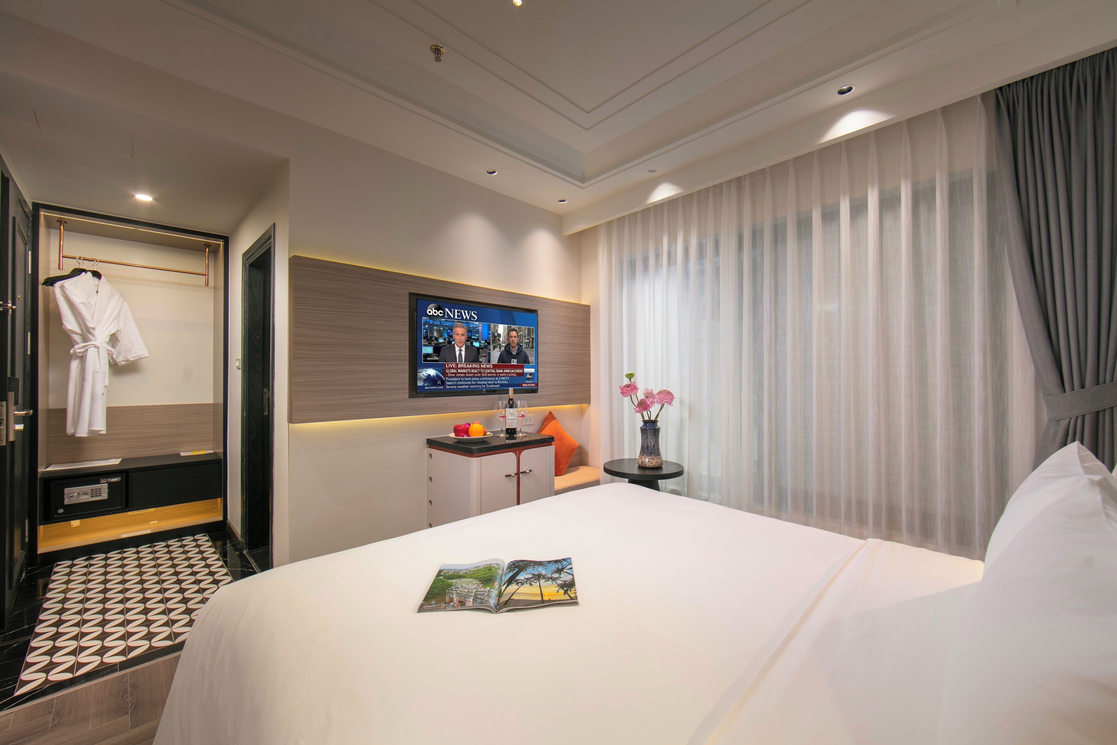 A hotel room with a bed, a TV mounted on the wall, a bedside table, a robe hanging on a door, and a window with curtains.
