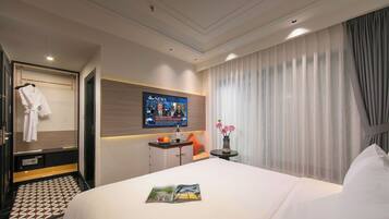 A hotel room with a bed, a TV mounted on the wall, a bedside table, a robe hanging on a door, and a window with curtains.
