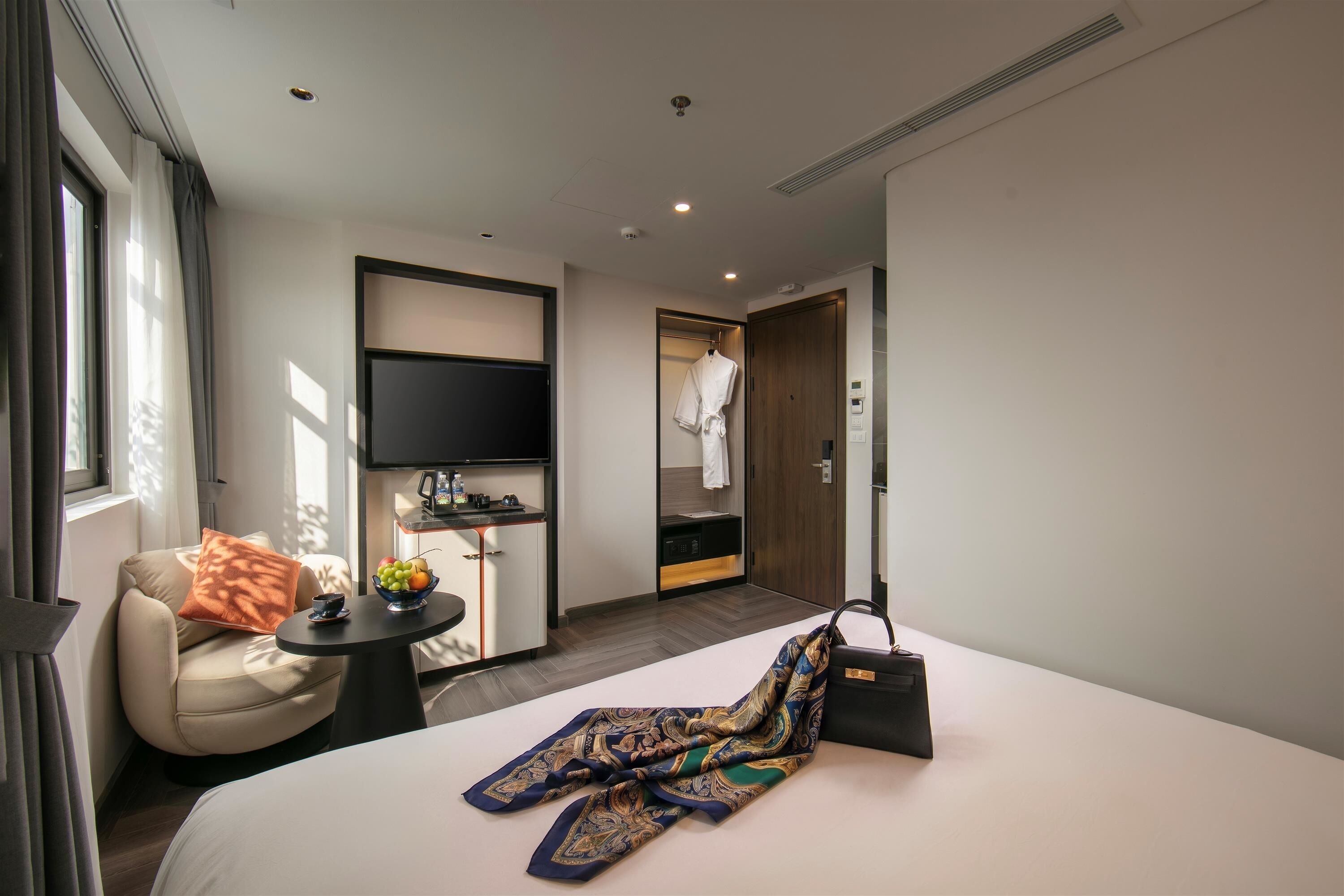 A hotel room with a bed, a small table with a fruit bowl, a chair, a robe hanging on a door, and a TV mounted on the wall.