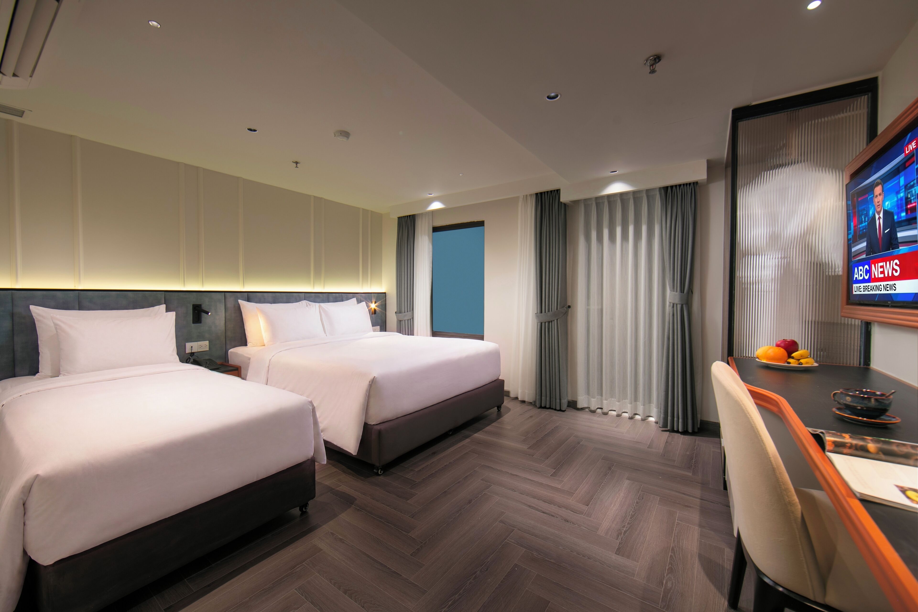Ein Hotelzimmer mit zwei Betten, einem Schreibtisch mit Stuhl, einem Fernseher und einem Fenster mit Vorhängen.