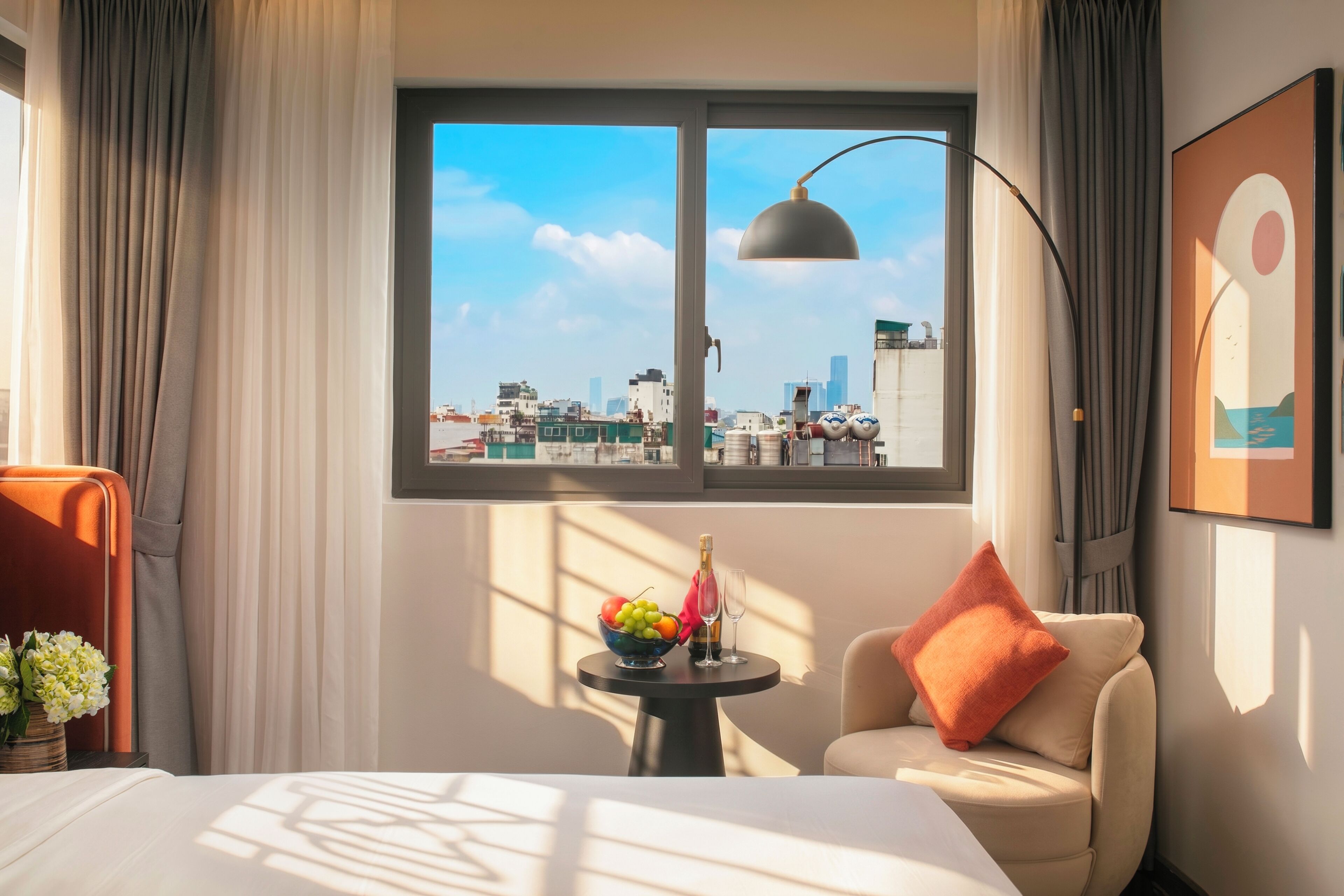 A hotel room with a bed, a small table with a fruit bowl and a bottle, a sofa with a cushion, a window with a city view, and a lamp.