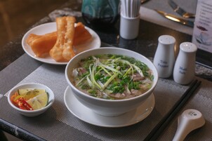 A bowl of soup with vegetables and meat, a plate of fried items, and a small bowl of sliced vegetables on a table.