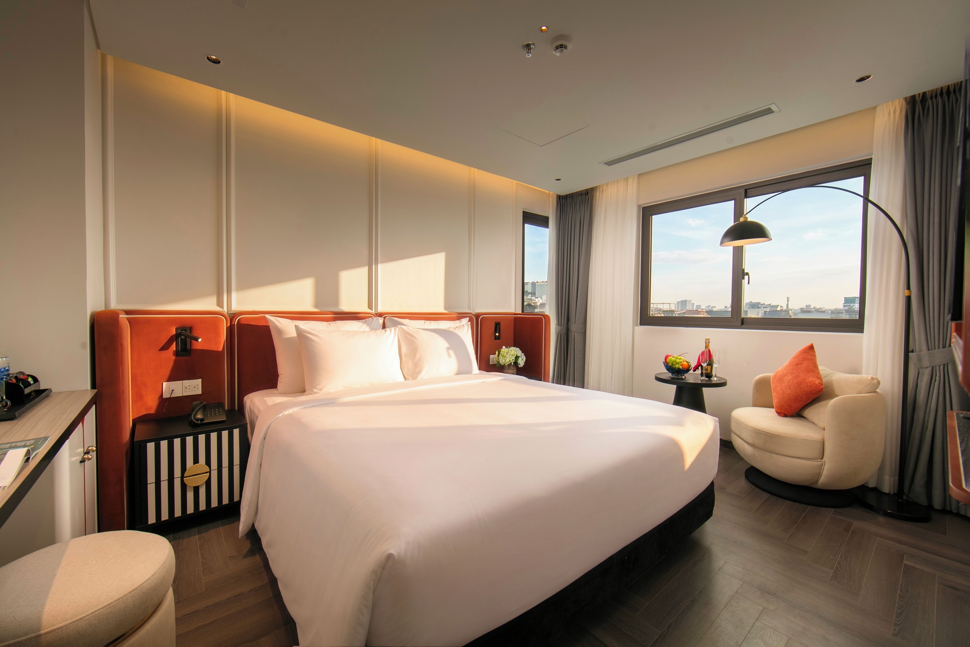 A hotel room with a large bed, a desk, a chair, a small table with a lamp, and a view of the city through large windows.