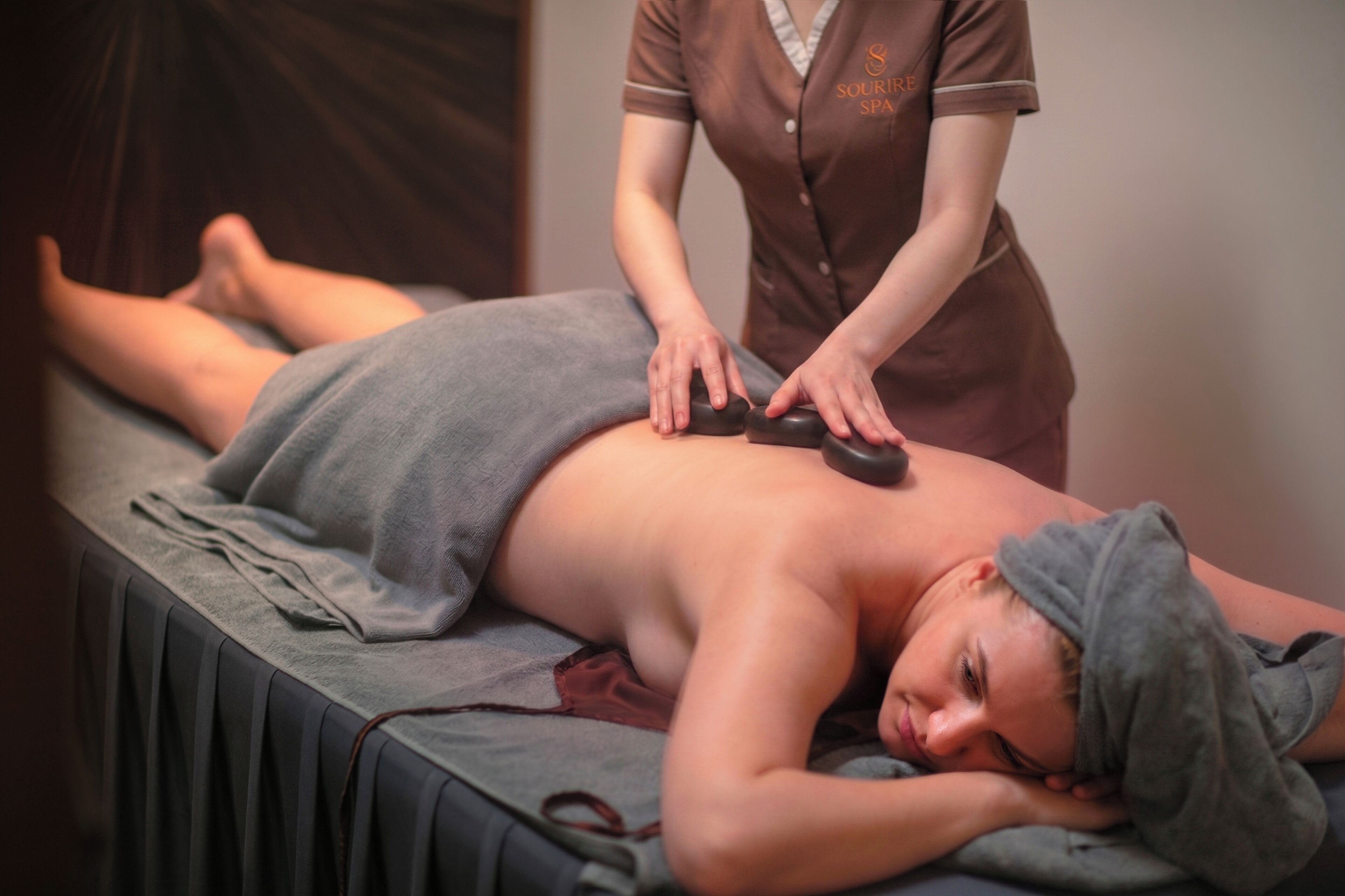 A person receiving a massage with a towel covering their lower body, a massage table, and a masseuse in a uniform.