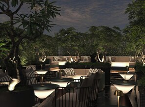 An outdoor seating area with round tables and cushioned benches, surrounded by lush greenery and trees. - May De Ville Lumiere Hotel (Hoan Kiem)