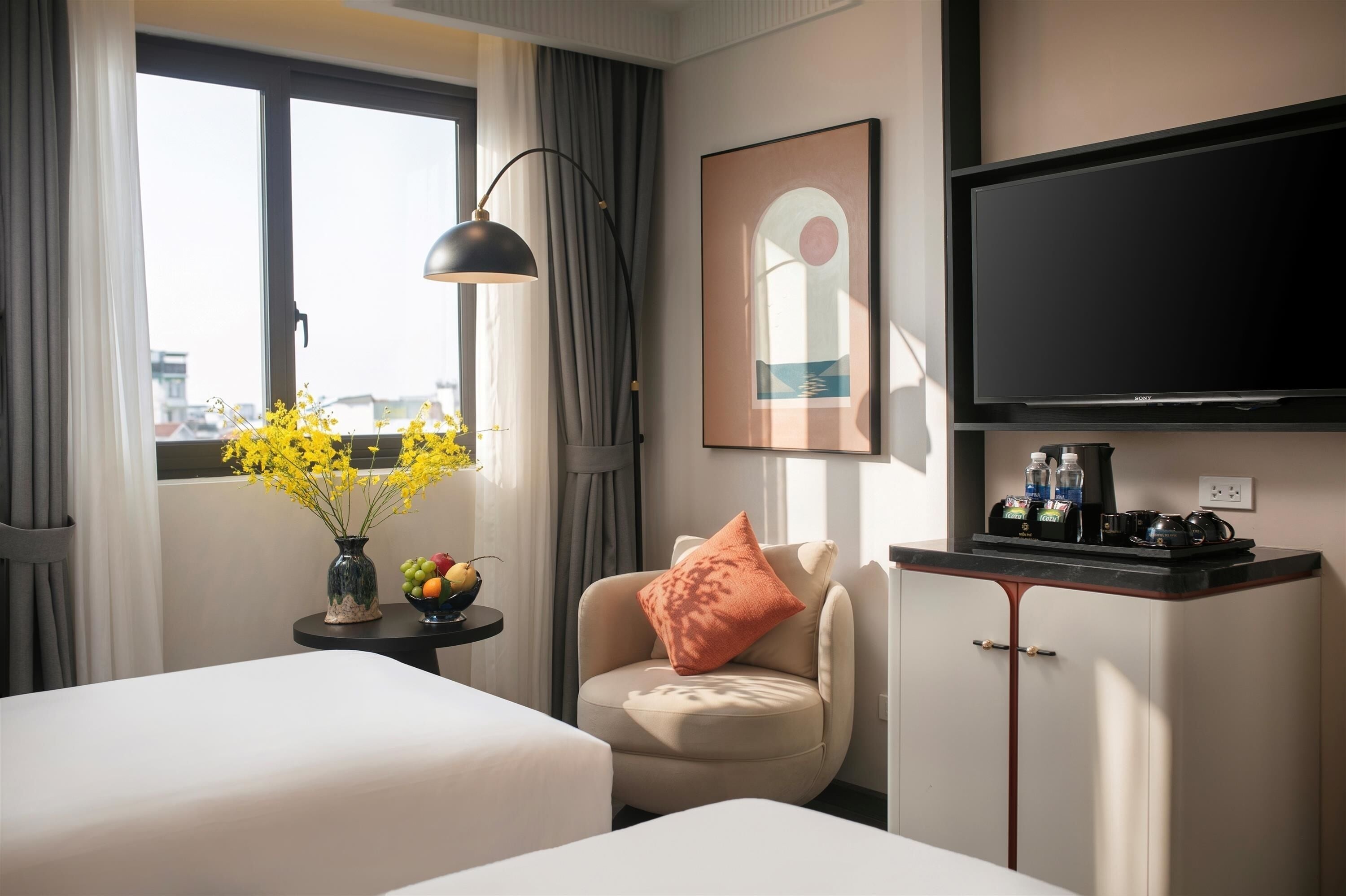 Habitació d'hotel amb dos llits, un escriptori amb una làmpada, una televisió, un quadre amb marc i una finestra amb cortines.