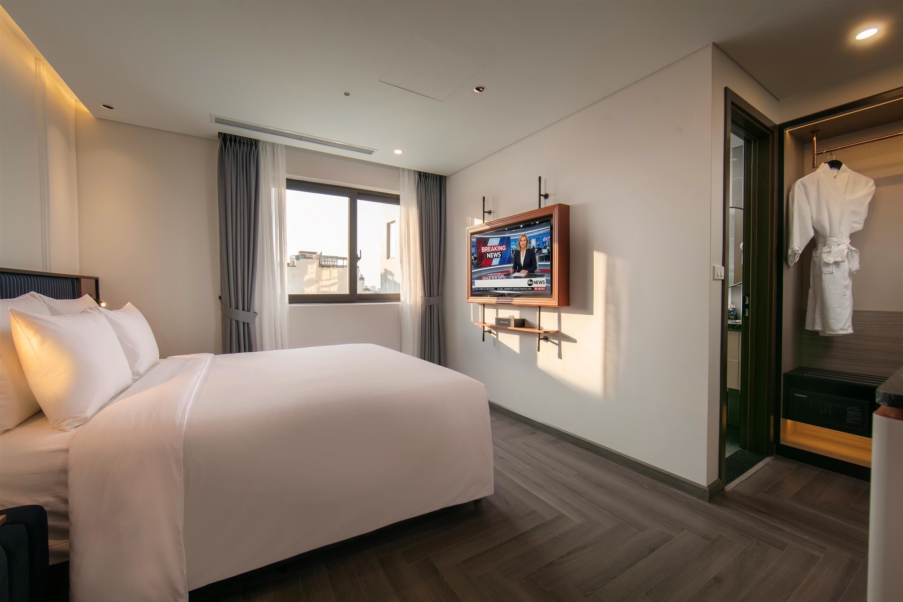 Une chambre d’hôtel avec un grand lit, un téléviseur fixé au mur, un peignoir suspendu à un porte-manteau et une vue sur la ville par la fenêtre.