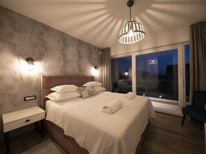 Et moderne hotelværelse med en stor seng, sengeborde, lamper og udsigt over byen om natten.
