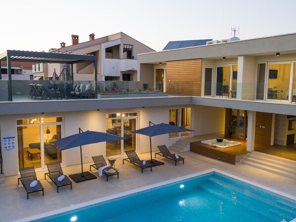 Et moderne hus med to etager, en swimmingpool, udendørs møbler og et spabad.