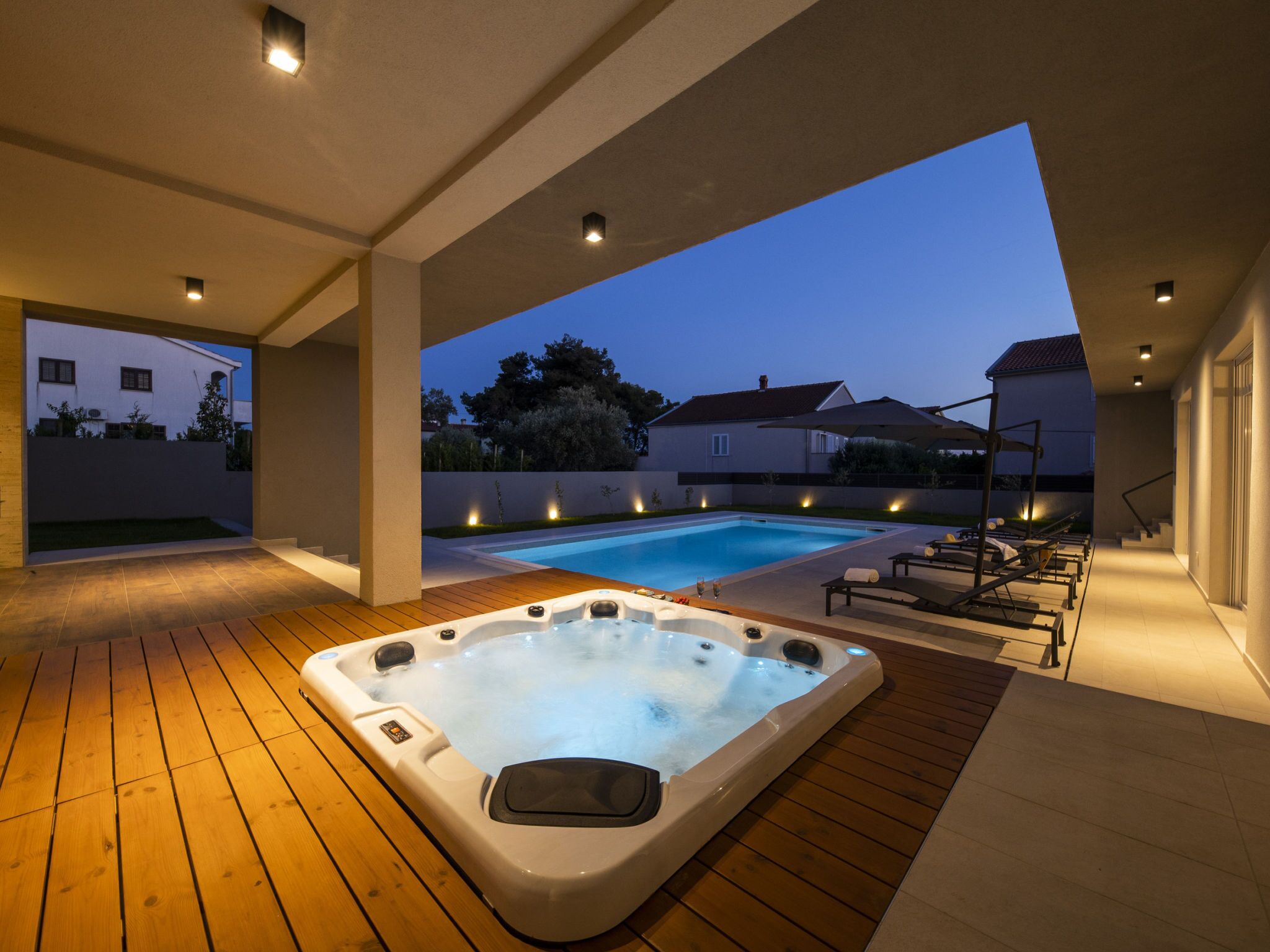 A hot tub with a wooden deck, a swimming pool, and lounge chairs in a residential setting.