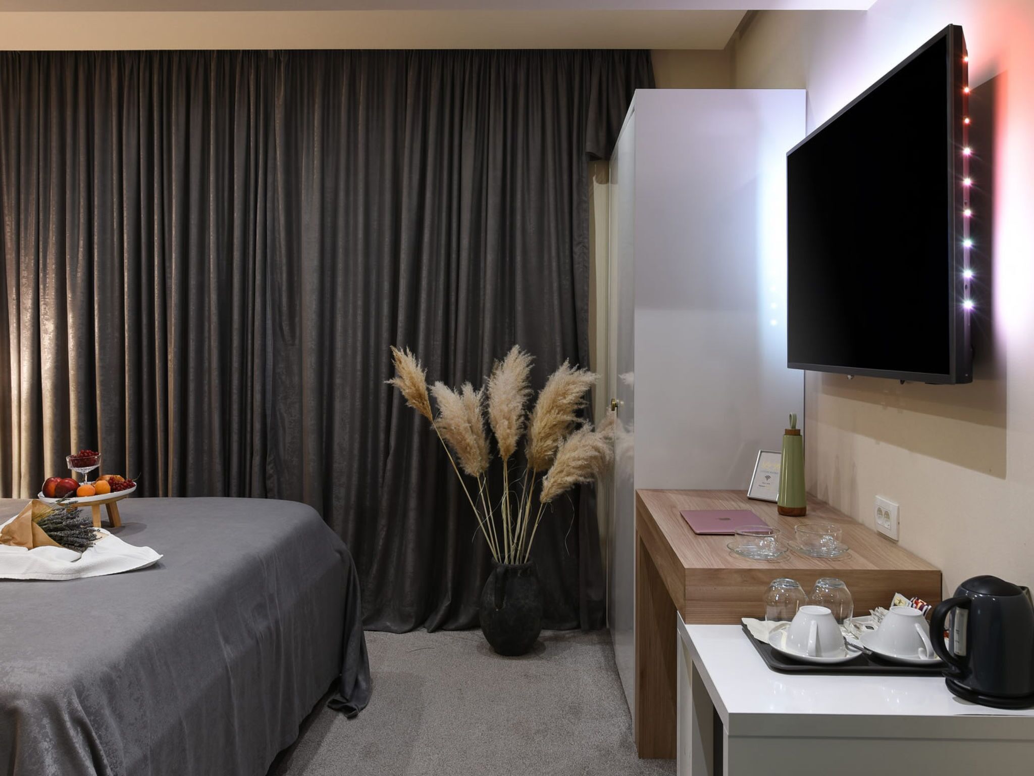 Une chambre d’hôtel avec un lit, un bureau doté d’un téléviseur à écran plat, un vase contenant des plumes d’herbe des pampas et une petite table avec un panier de fruits et des verres.