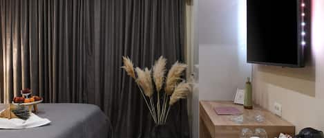 A hotel room with a bed, a desk with a flat-screen TV, a vase with pampas grass, and a small table with a fruit bowl and glasses.