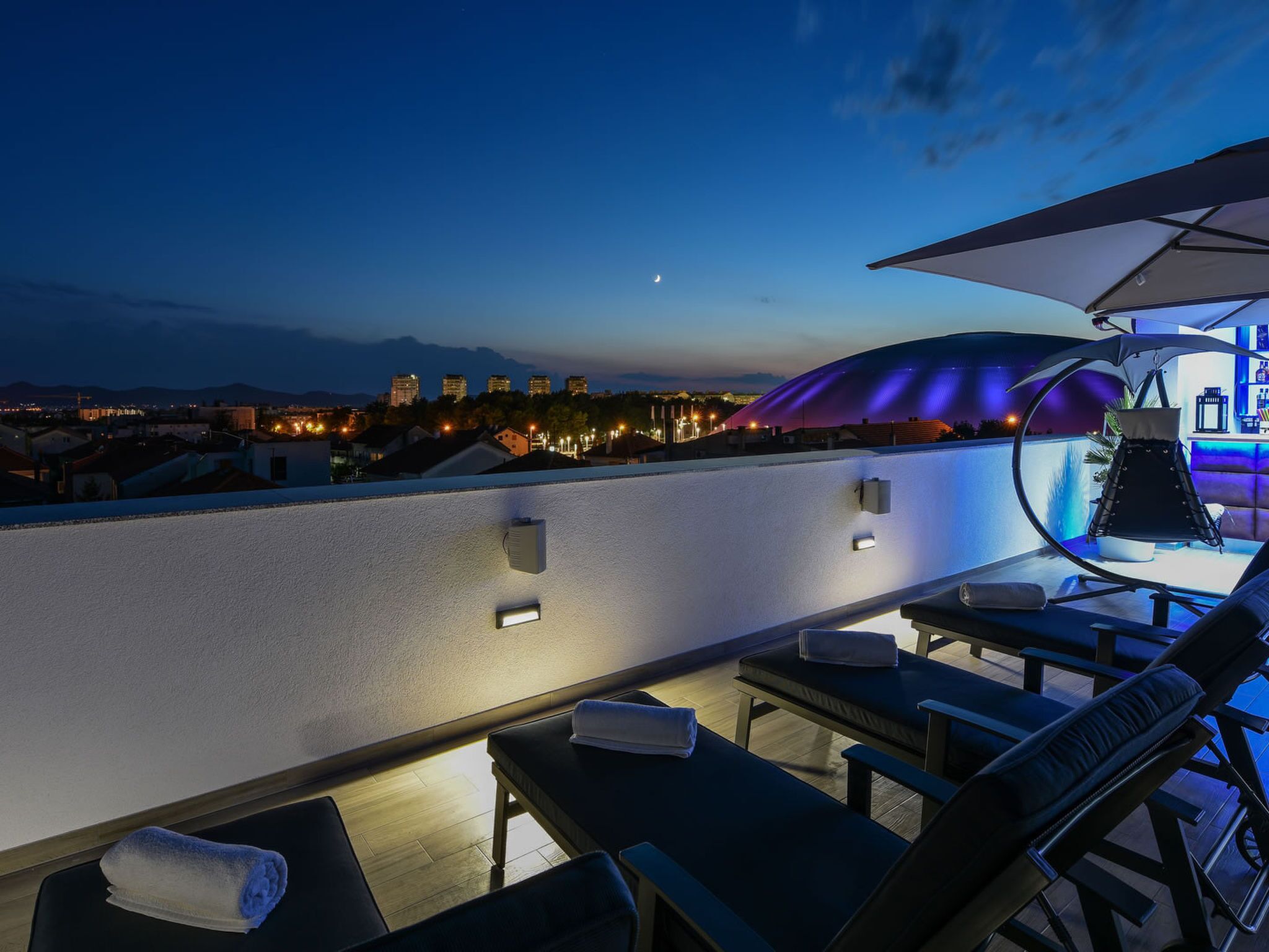 A rooftop terrace with lounge chairs, a bar area, and a cityscape view at dusk.