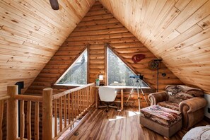 Eine Holzhütte mit geneigtem Dach, zwei Fenstern, einem Schreibtisch, einem Stuhl und einer Couch.