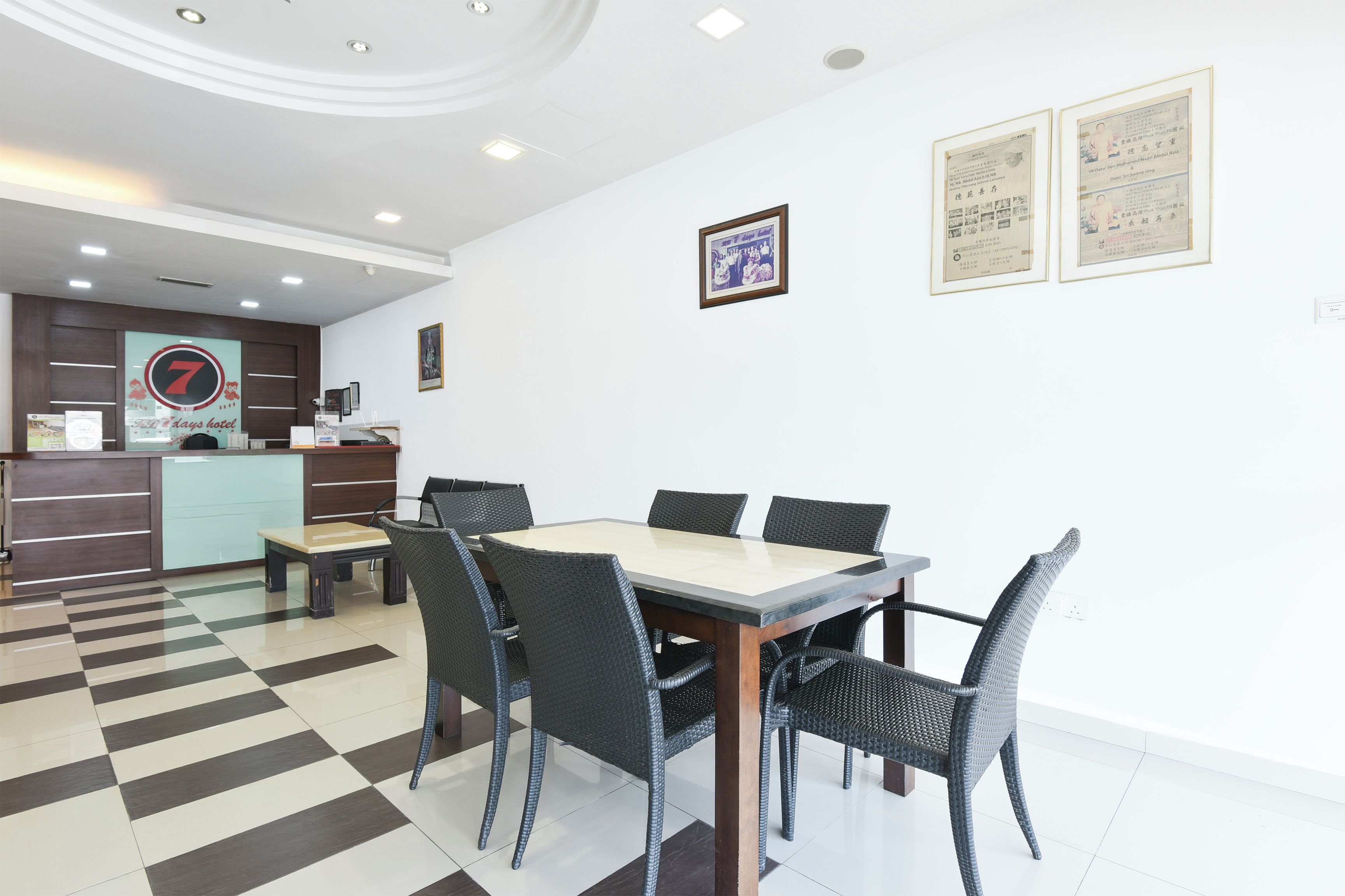 A hotel lobby with a check-in counter, a reception desk, a dining area with a table and chairs, and framed certificates on the wall.