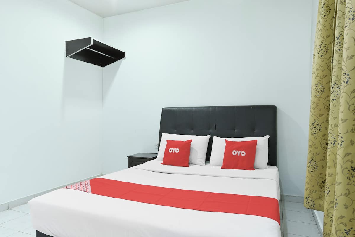 Una habitación de hotel con una cama, dos almohadas con la marca 'OYO', un cabecero negro, una mesita de noche y un estante montado en la pared.