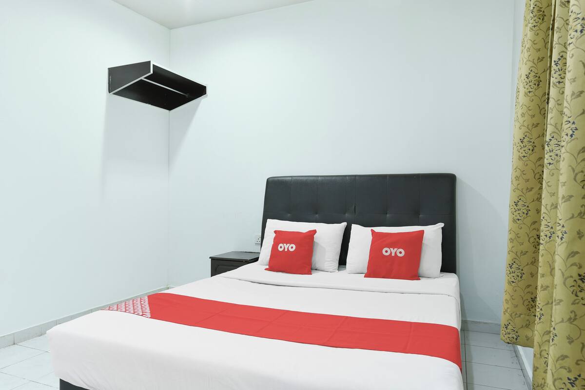A hotel room with a bed, two pillows with 'OYO' branding, a black headboard, a nightstand, and a wall-mounted shelf.