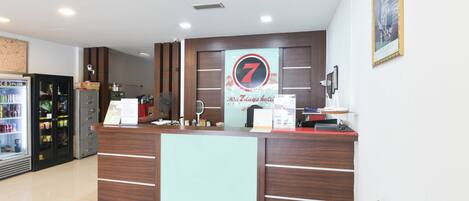 Ein Hotelrezeptionstresen mit einem '7'-Logo, einem Kühlschrank und einer Theke mit verschiedenen Artikeln.