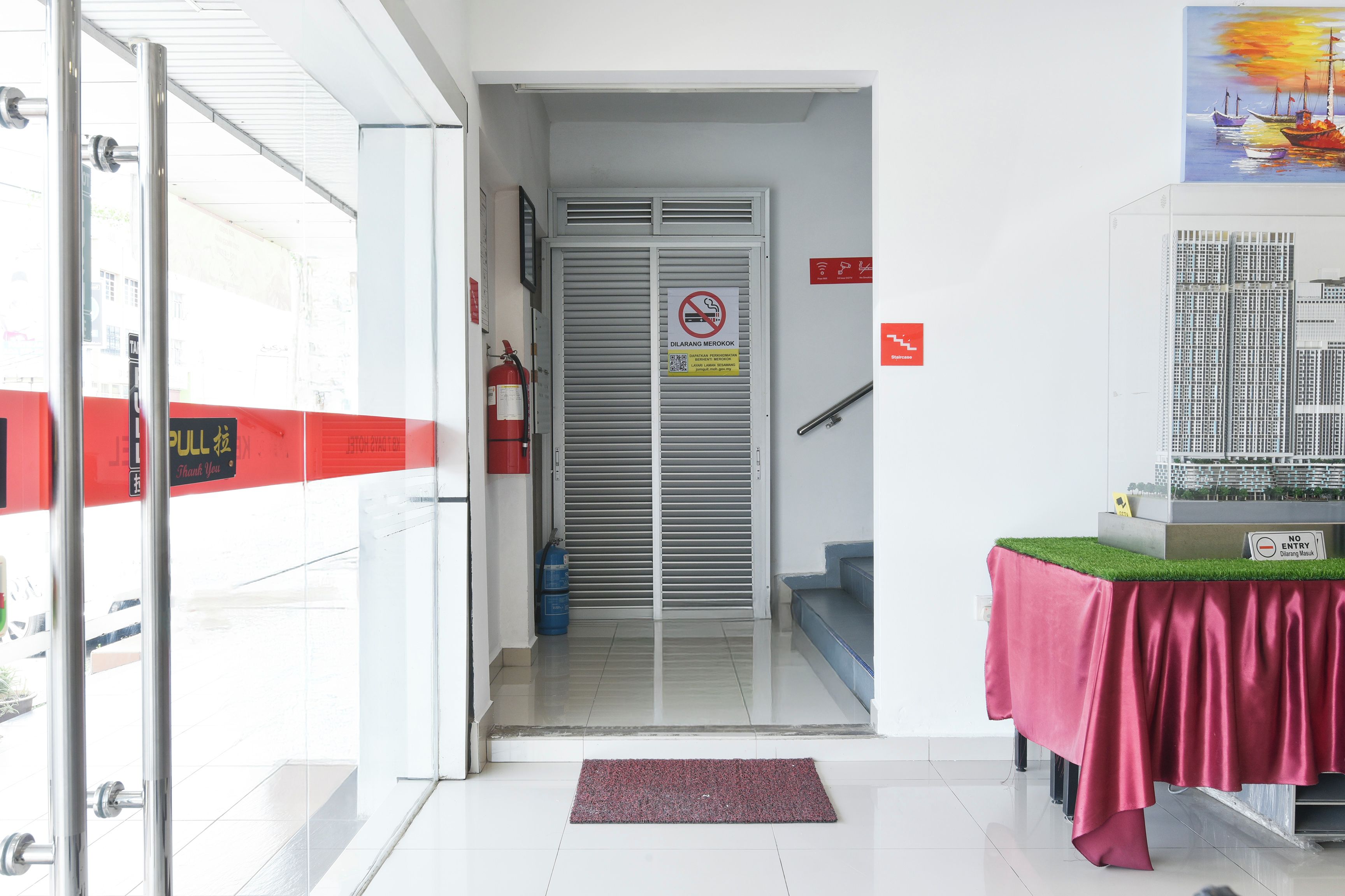 Entrada de un hotel con extintor, un cartel de prohibido fumar y puerta de cristal que da a una escalera.